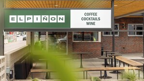 An outdoor seating area with tables and benches, a sign for Elpinon Coffee, Cocktails, and Wine, and a brick wall in the background. - Highway Daydream | La Vista Motel | 106 (Denver)