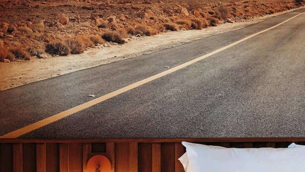 A room with a bed, a nightstand with a lamp, and a view of a desert road. - Highway Daydream | La Vista Motel | 106 (Denver)