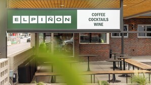 Un área con mesas y bancos para disfrutar al aire libre, un letrero que anuncia Elpinon Coffee, Cocktails y Wine, y un muro de ladrillo al fondo.