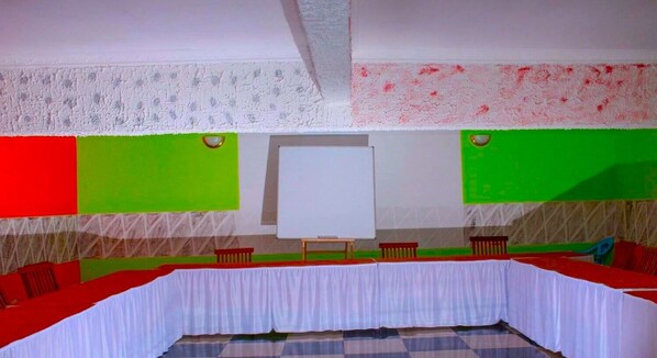 Un salón de conferencias con una pizarra, una mesa larga con manteles rojo y blanco, y paredes verdes.