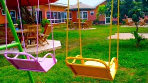 A swing set with two swings, a patio area with wooden furniture, and a building with large windows in the background. - kuwetu resort and conference (Muthara)