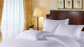 A neatly made bed with white bedding, a wooden headboard, and a bedside table with a lamp and a plant. - kuwetu resort and conference (Muthara)