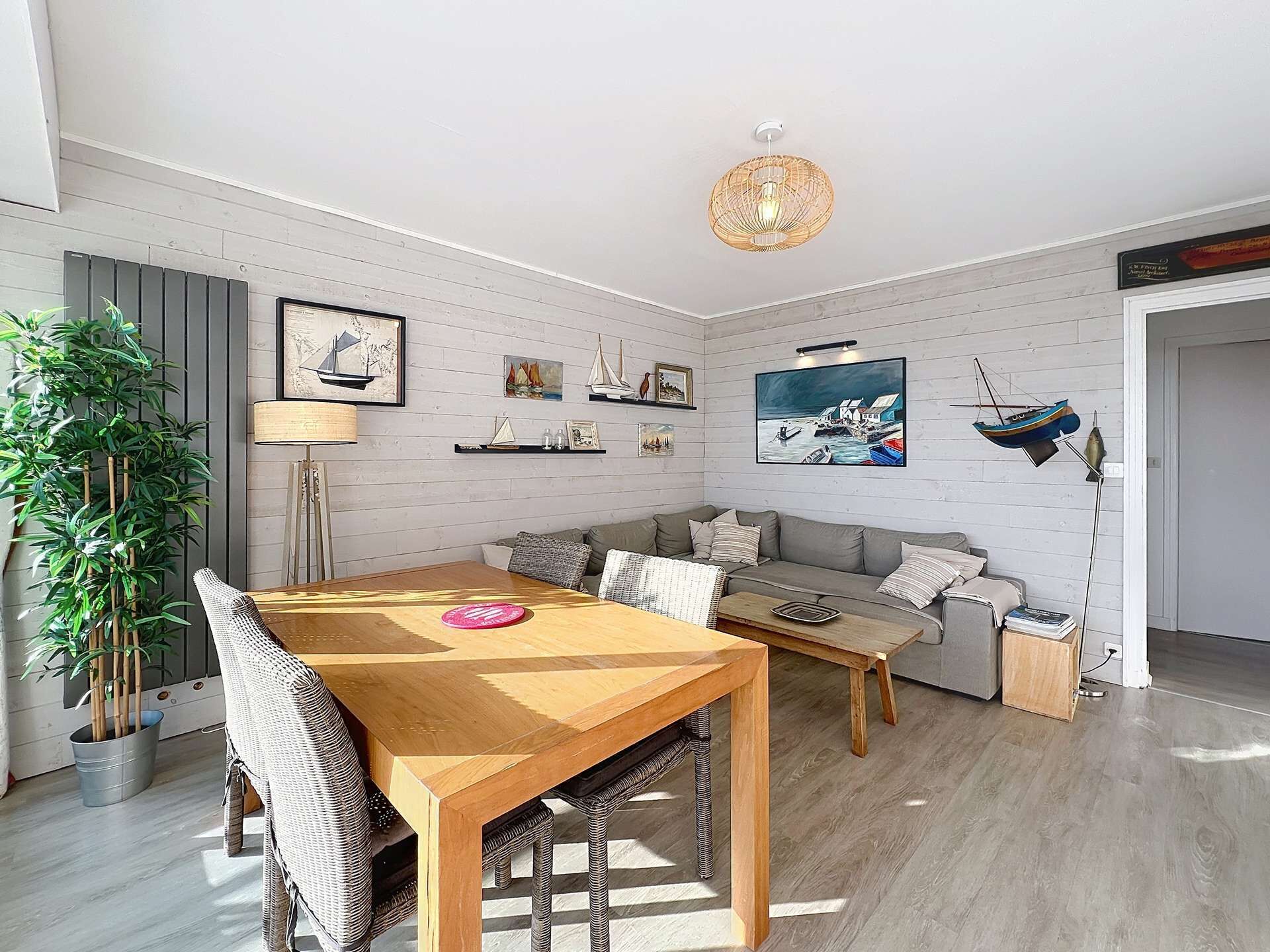 A modern living room with a wooden dining table, grey sofa, and a large potted plant.