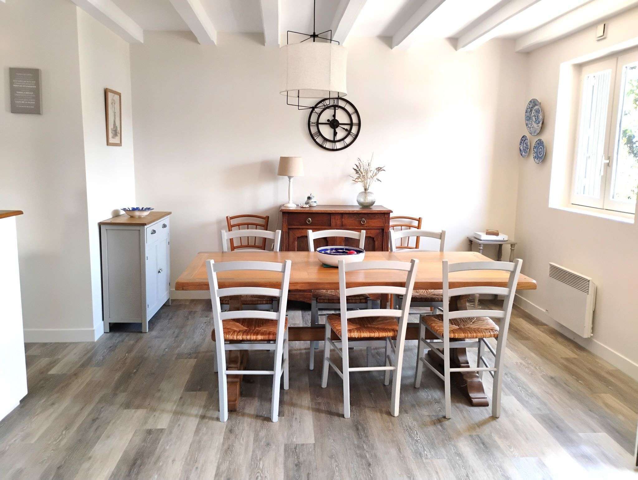 Un coin repas avec une table en bois et des chaises blanches, une horloge au mur et un petit meuble sur le côté gauche.