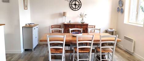 Un coin repas avec une table en bois et des chaises blanches, une horloge au mur et un petit meuble sur le côté gauche.