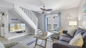 Una sala de estar con televisor de pantalla plana, ventilador de techo, escalera y vista a la playa.