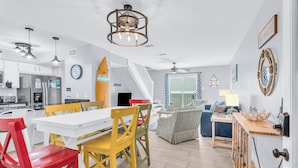 Una cocina moderna con armarios blancos, un área de comedor con sillas coloridas, una escalera y una sala de estar con un sofá y una mesa de centro.