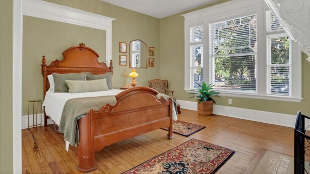 A bedroom with a wooden bed, a nightstand, a lamp, a chair, a window with blinds, and a potted plant. - Newly Renovated Victorian APT, Steps from Forsyth Park (Savannah)
