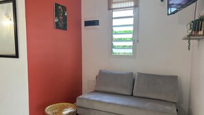 Un salon avec un canapé gris, une petite table ronde, une fenêtre avec des stores, un téléviseur fixé au mur et un tableau encadré.