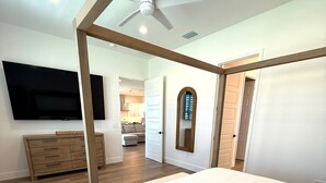 Un dormitorio con una cama grande, suelo de madera, ventilador de techo, televisor y vistas al salón.
