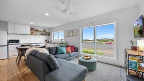 A modern kitchen with white cabinets, a dining area with a grey sofa and ottoman, and large windows offering a view of a road and greenery. - The Bungalow - Your Stylish Warrnambool Retreat (Warrnambool)