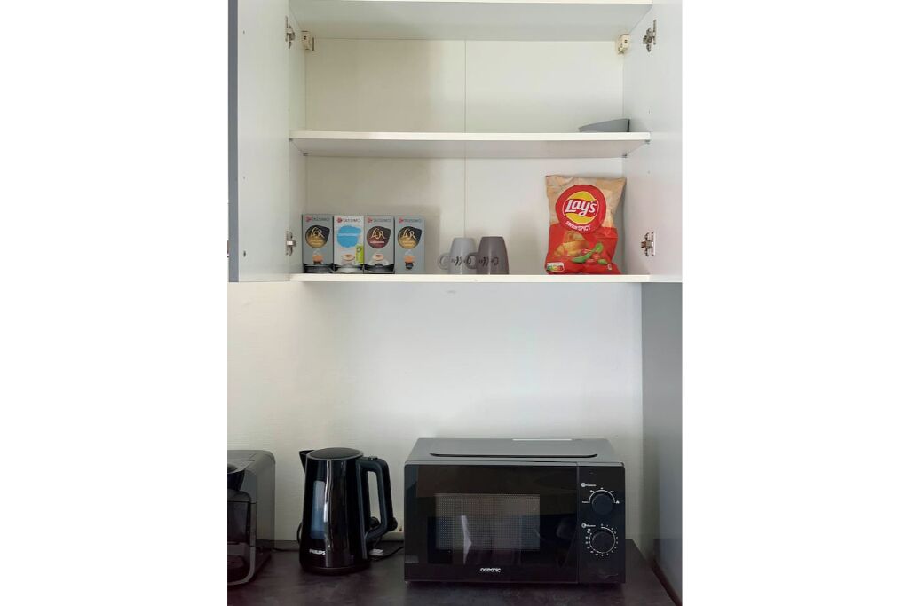 Een keukenhoek met een magnetron, koffiezetapparaat en een plank met snacks en koffiecups.