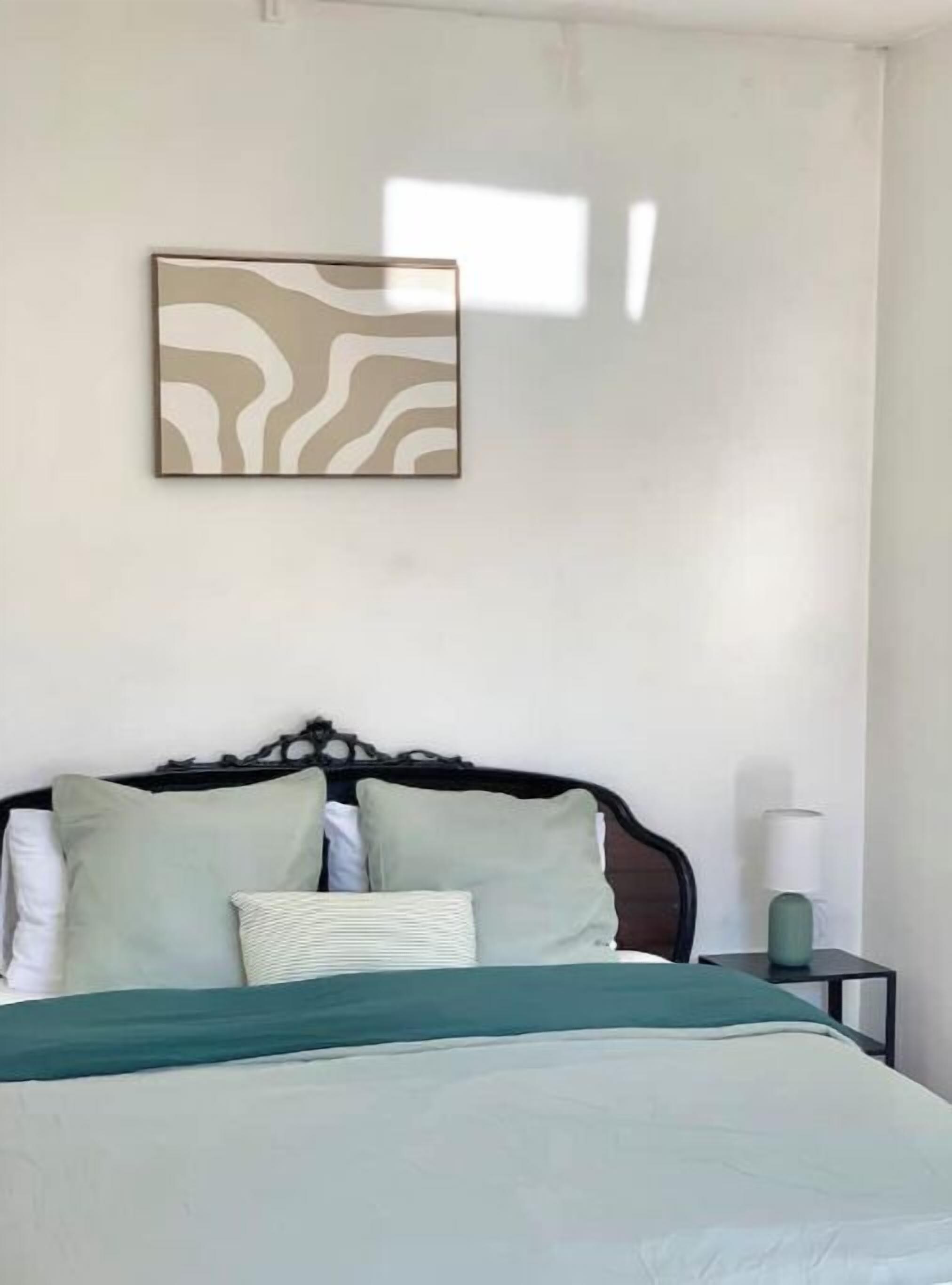 Un dormitorio con cama, almohadas, una mesita de noche y una obra de arte en la pared.