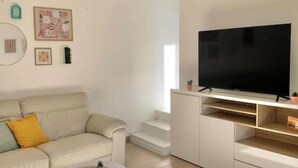A modern living room with a beige sofa, a coffee table, a flat-screen TV on a white stand, and a staircase leading to an upper level. - Charming house with terrace near Paris (Viry-Châtillon)