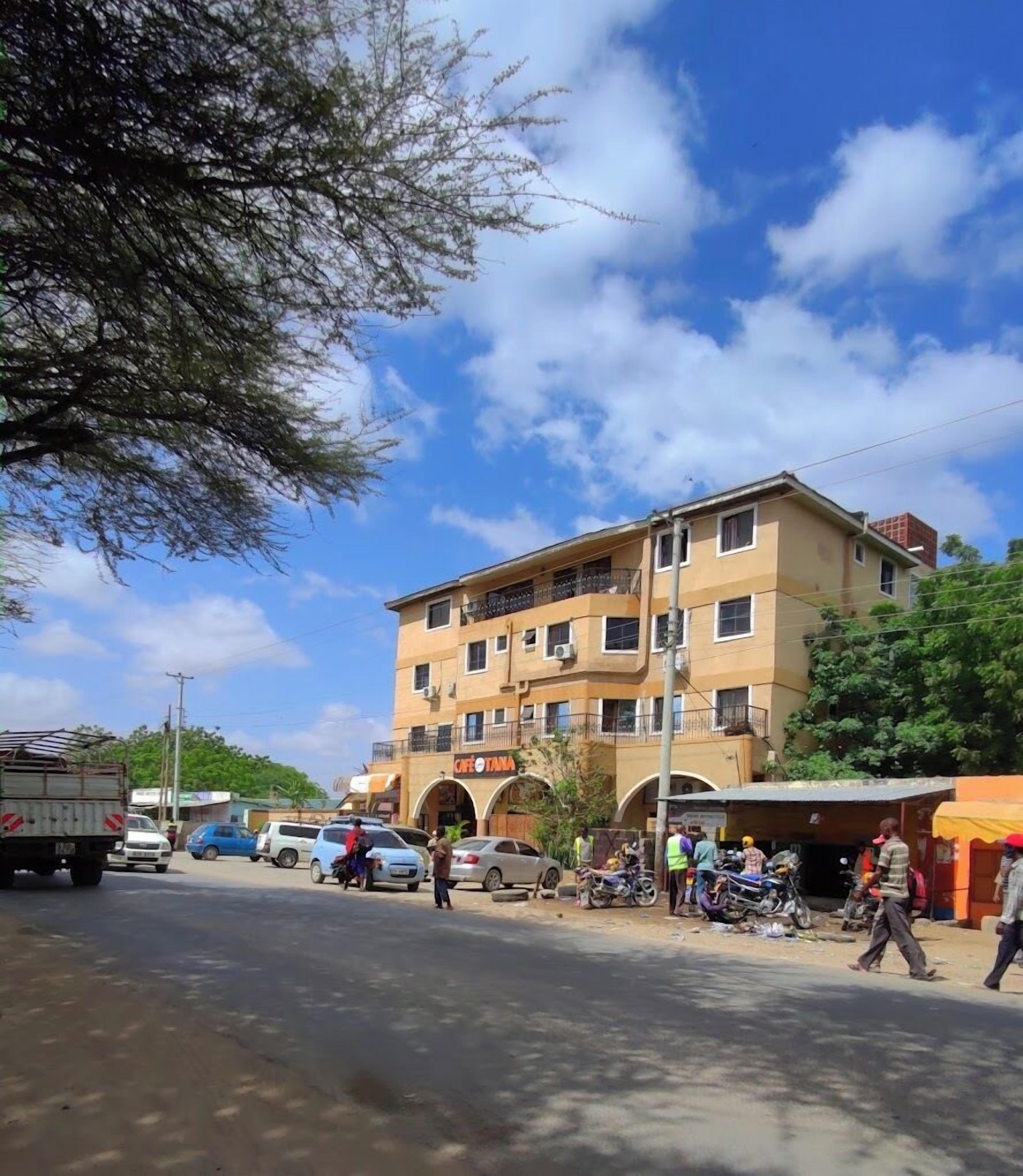 A multi-story building with a cafe named "Café Tana" on the ground floor. There are several parked cars and motorcycles, and a few people are visible on the street.