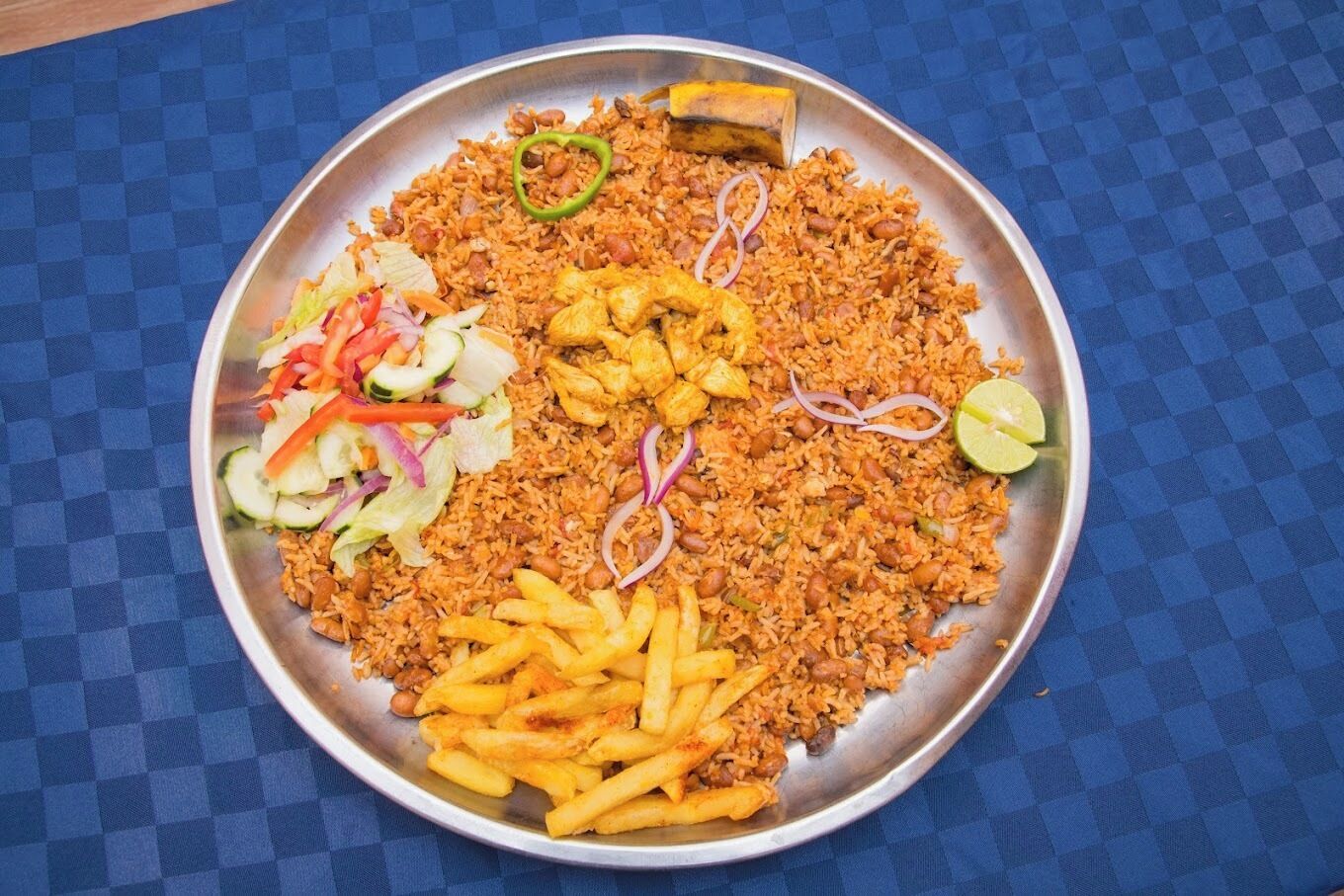 Un plato grande de metal con diversos alimentos, como arroz, pollo y papas fritas, acompañado de una ensalada y gajos de limón.