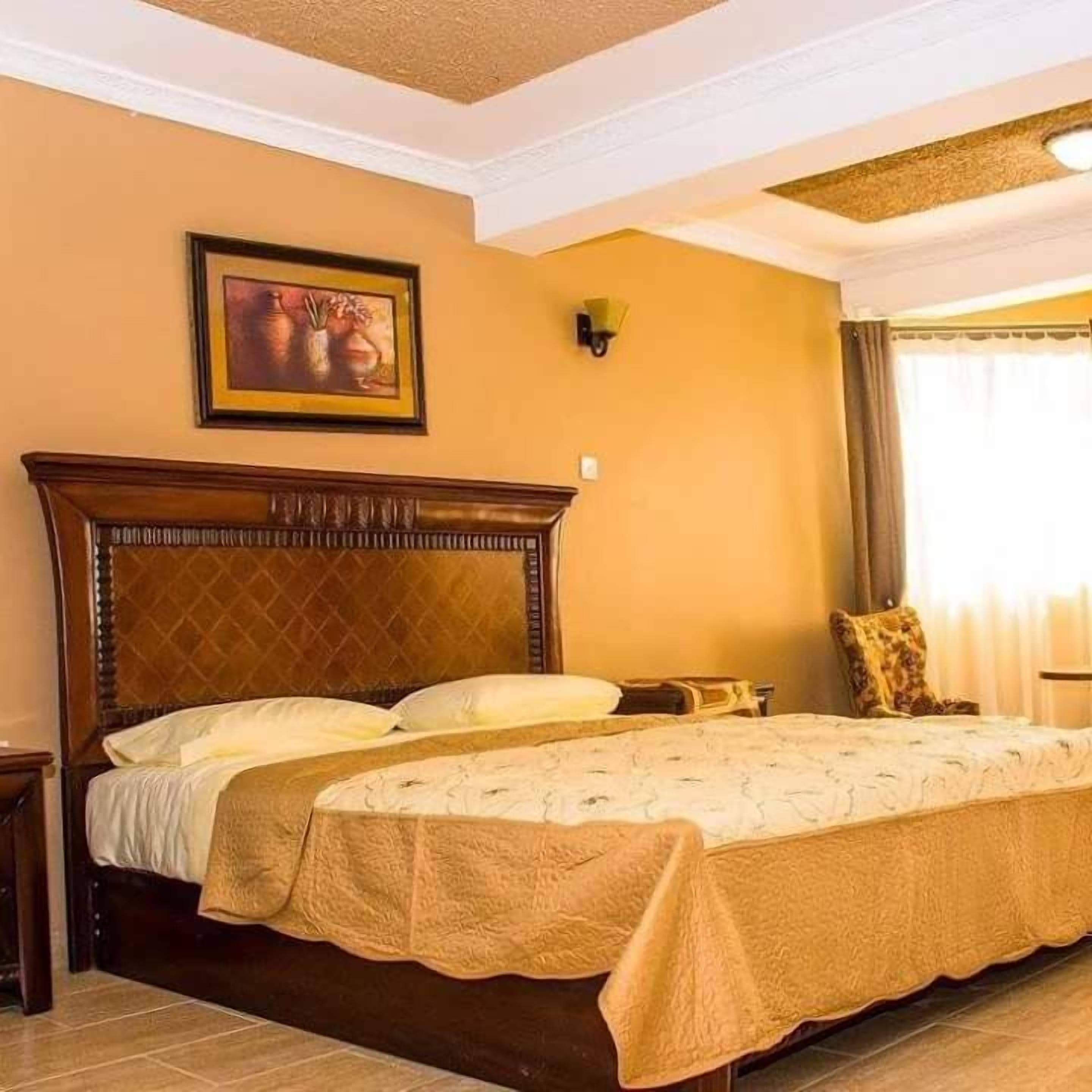 A hotel room with a large bed, a wooden headboard, a painting on the wall, and a window with curtains.