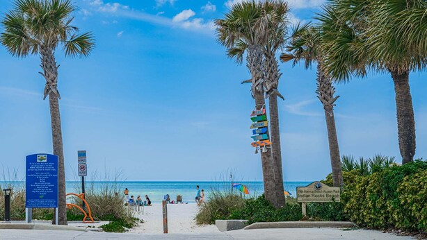 A beach entrance with palm trees, a sign for 19th Avenue Beach Access Park, and a beach with people and umbrellas.