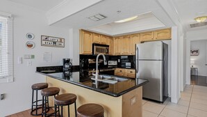 A modern kitchen with wooden cabinets, a stainless steel refrigerator, a built-in microwave, a coffee maker, and a sink. There are four stools at the kitchen island.