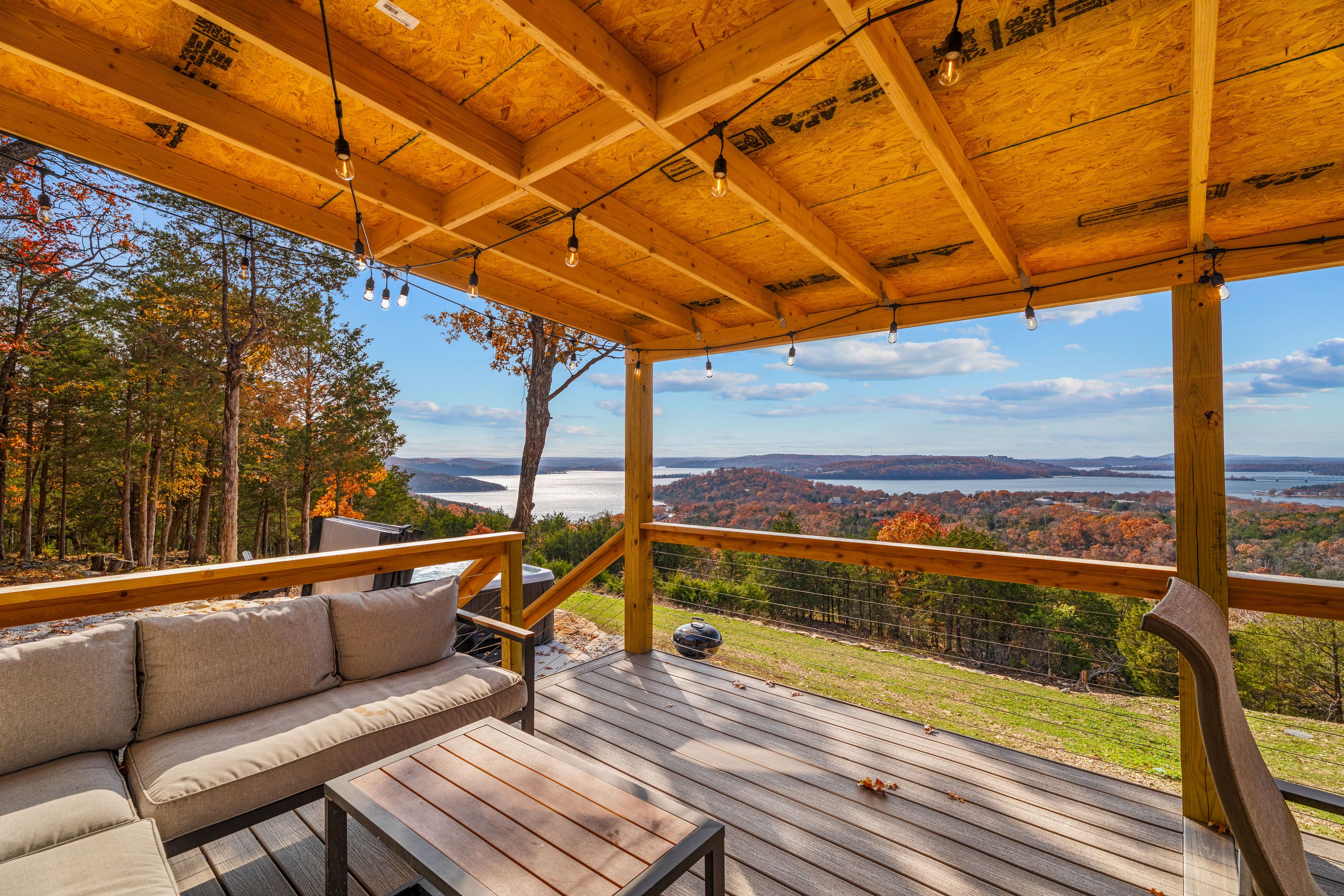 Une terrasse en bois avec un canapé et un jacuzzi, offrant une vue panoramique sur un paysage verdoyant, avec des arbres et une étendue d’eau.