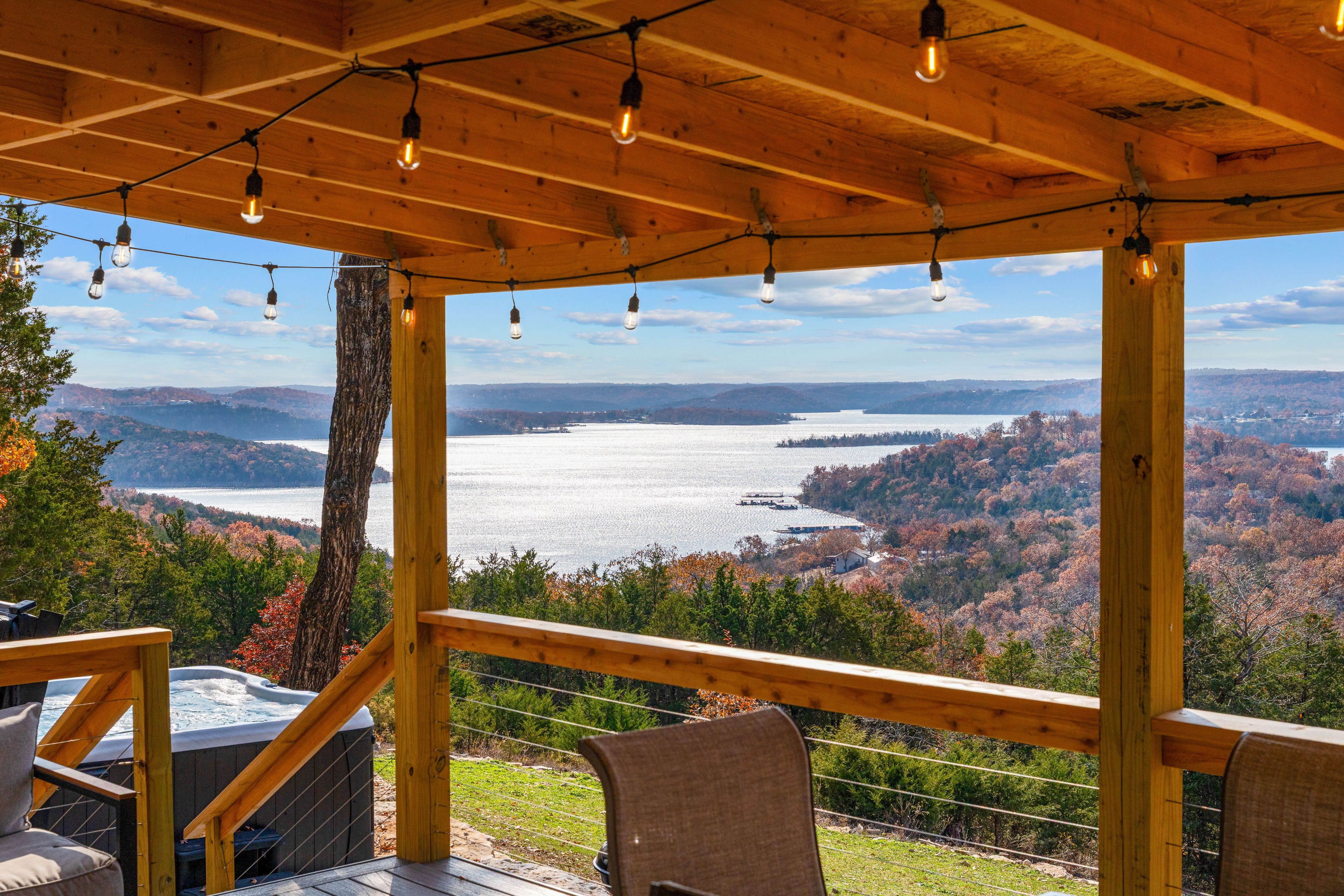 Une terrasse en bois avec un jacuzzi, des chaises et une vue sur un lac entouré d’arbres.
