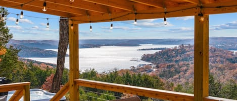 A wooden deck with a hot tub, chairs, and a view of a lake surrounded by trees.