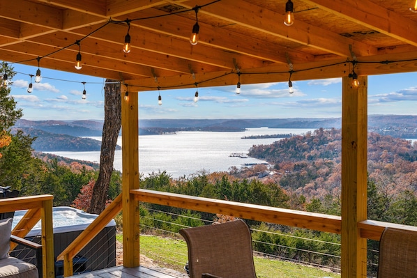 A wooden deck with a hot tub, chairs, and a view of a lake surrounded by trees. - Tucked Away Tiny Home Retreat | Sleeps 6 | Incredible Views (Reeds Spring)