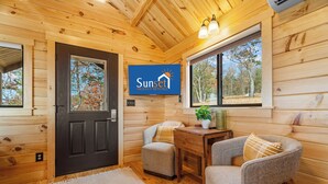 L’intérieur d’un chalet accueillant, avec une porte en bois, un téléviseur diffusant une émission intitulée « Sunset Nightly Rentals », une petite table ornée d’une plante et deux fauteuils rembourrés.