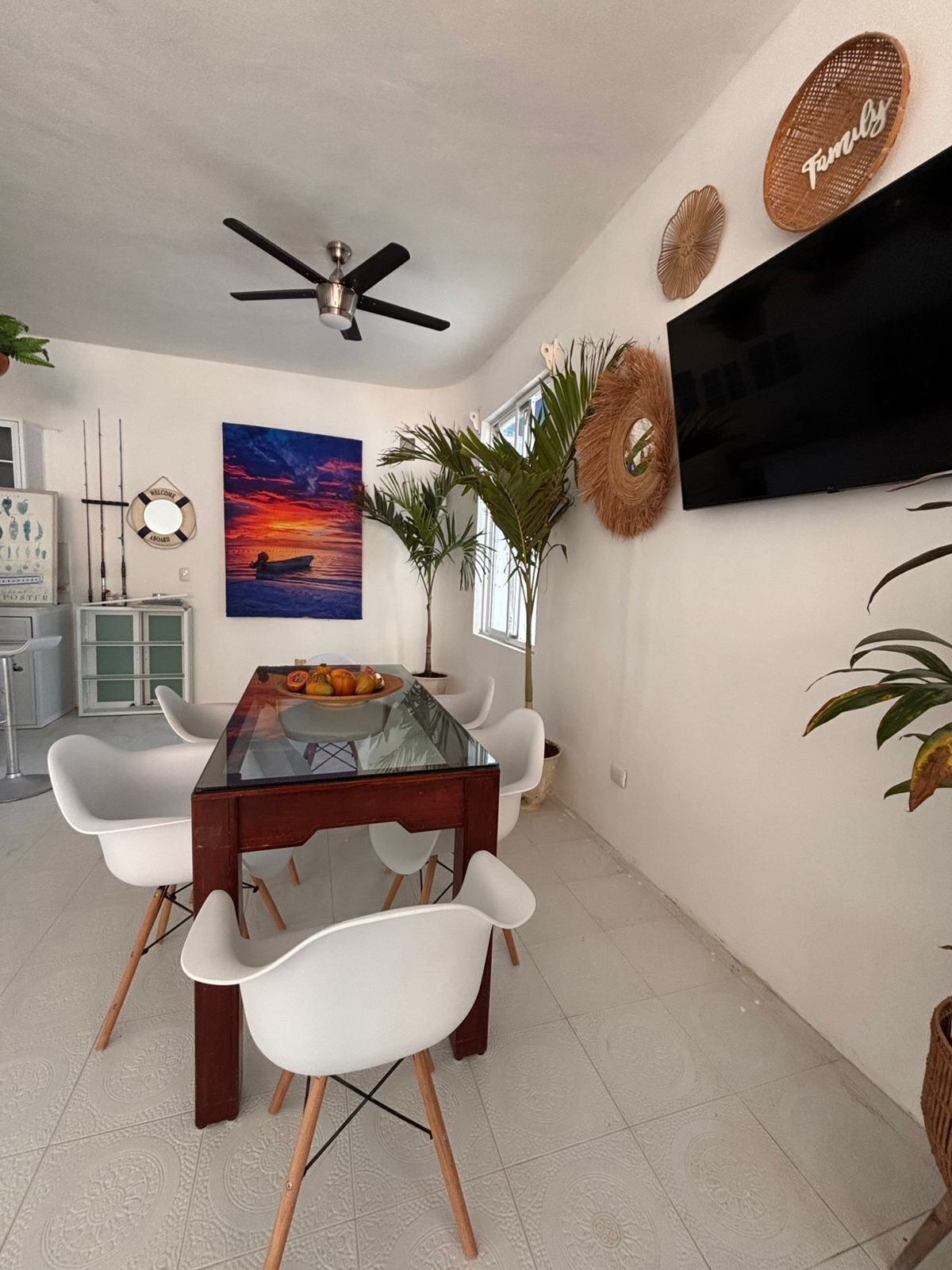 Un comedor con una mesa de vidrio, sillas blancas, un ventilador de techo, un televisor montado en la pared y un cuadro de un atardecer.