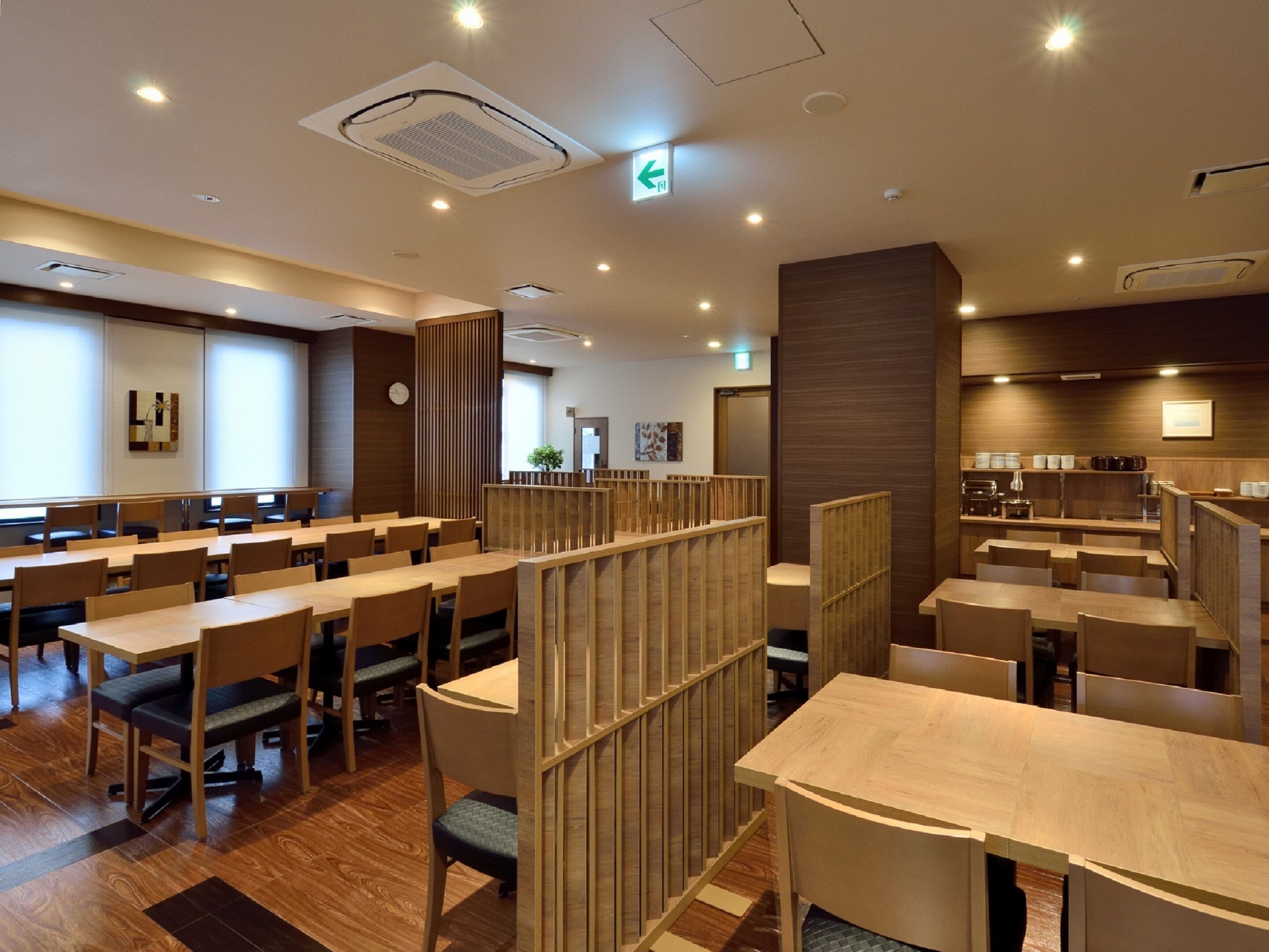 A modern hotel dining area with multiple tables and booths, wooden partitions, and a kitchen area in the background.