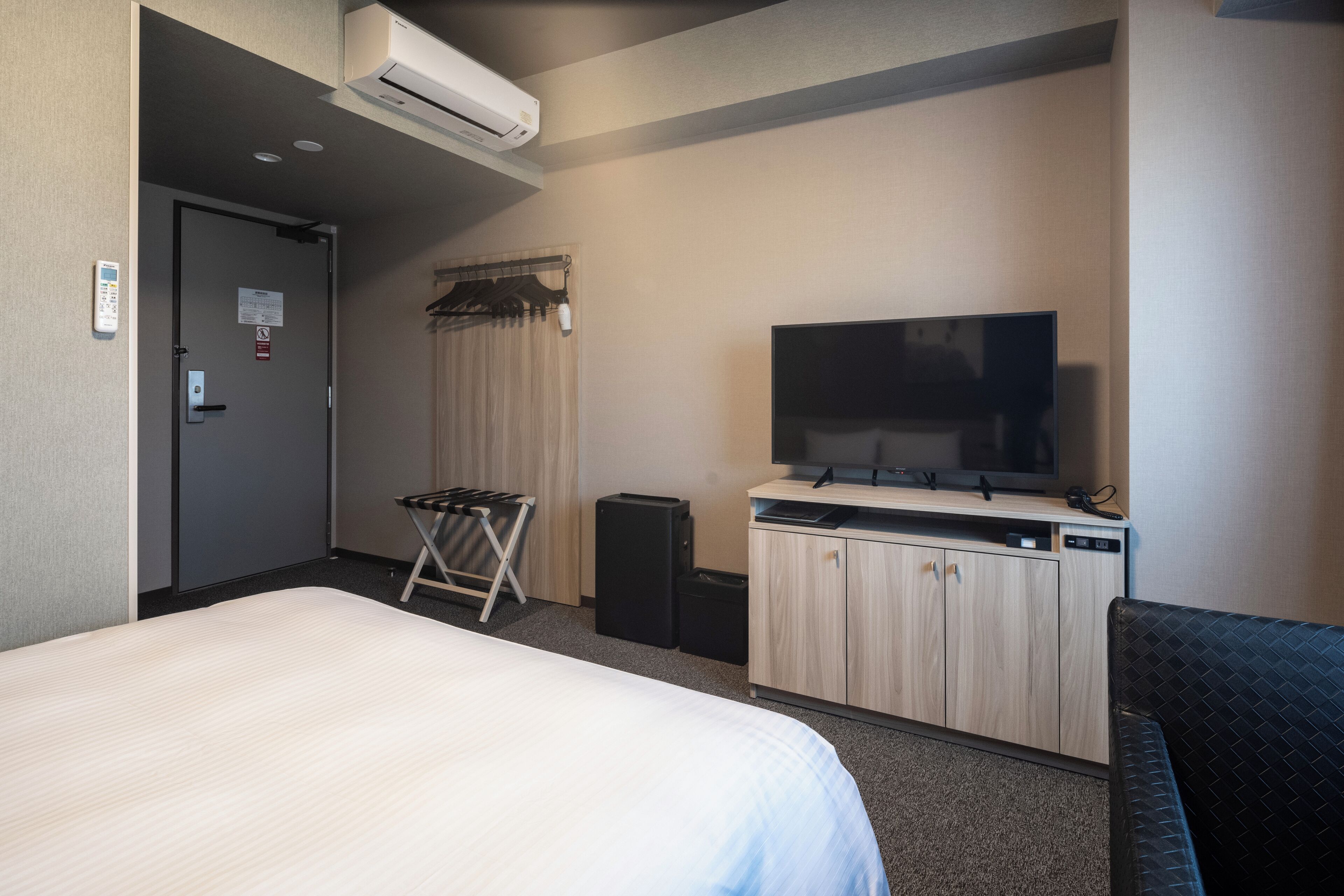 A hotel room with a bed, a TV, a small kitchenette, a wardrobe, and a door with a control panel.