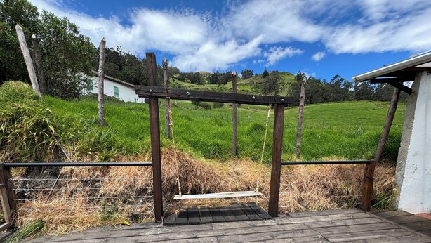 Um deck de madeira com estrutura de pérgola, cercado por um campo gramado e um edifício branco ao fundo.