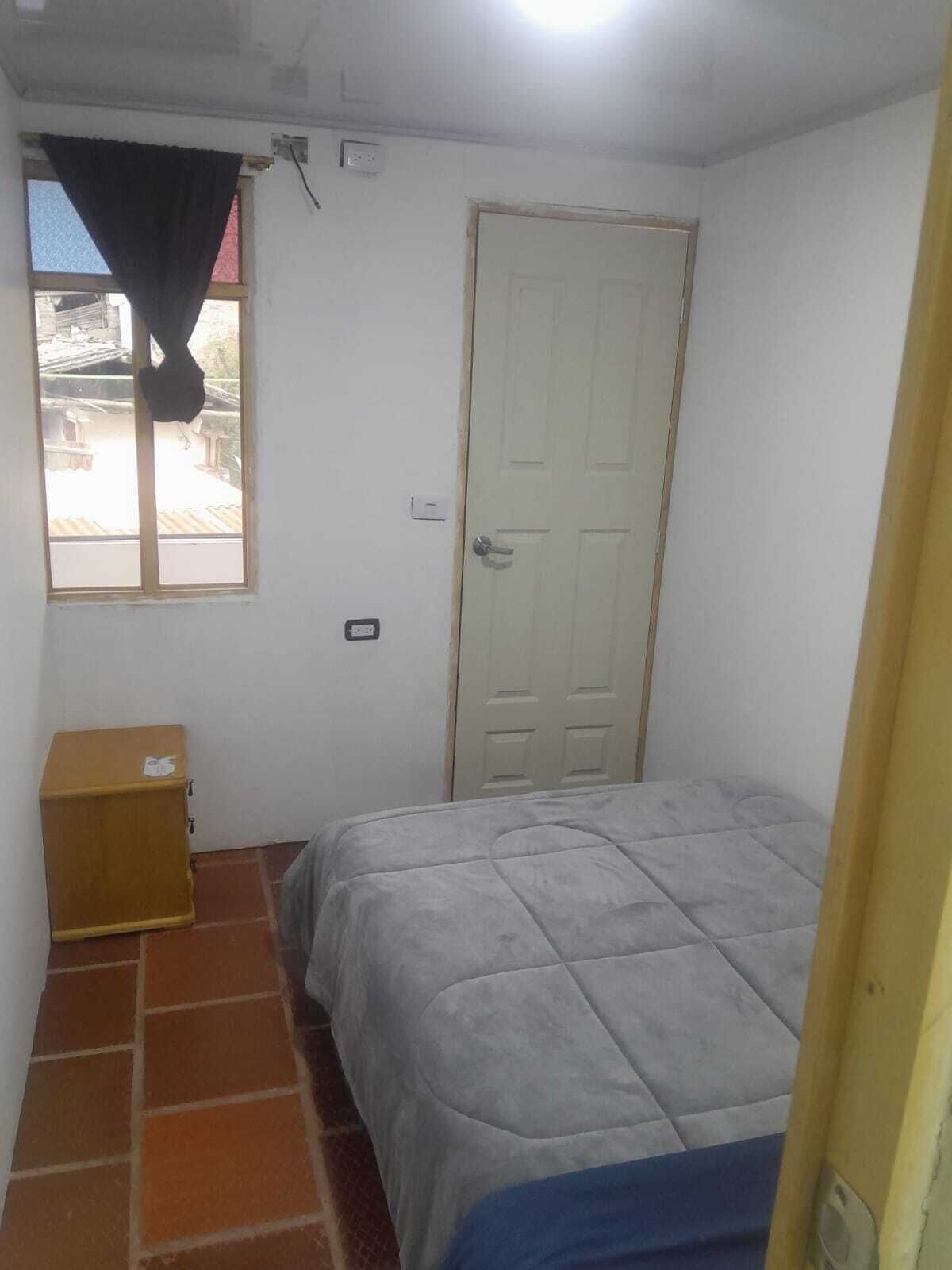 A small, simple room with a bed, a window with a curtain, a door, and a small wooden cabinet.