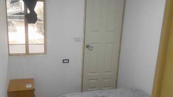 A small, simple room with a bed, a window with a curtain, a door, and a small wooden cabinet.