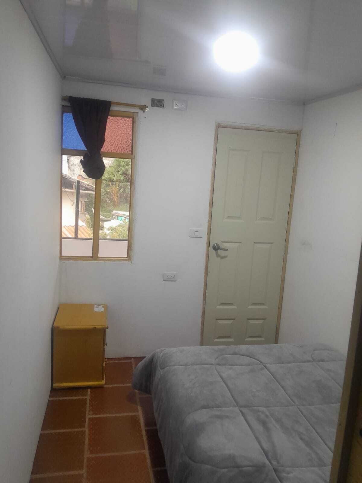 Una habitación pequeña y sencilla con cama, ventana con cortina, puerta y un mueble pequeño.