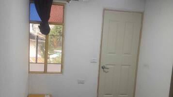 Una habitación pequeña y sencilla con cama, ventana con cortina, puerta y un mueble pequeño.