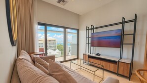 Un salon moderne doté d’une grande fenêtre, d’un canapé, d’un téléviseur fixé sur un cadre en métal noir et d’un balcon offrant une vue sur les bâtiments et la mer.