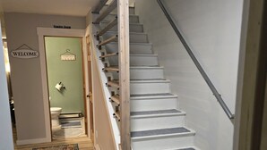 Un escalier avec une rampe en bois et un palier recouvert de moquette. On peut voir au fond une pancarte de bienvenue et une pancarte indiquant l’emplacement de la buanderie.