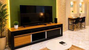 Ein modernes Wohnzimmer mit einem großen Flachbildfernseher, einem Holzkonsolentisch, einem runden Couchtisch sowie einem Essbereich mit Stühlen und einem kleinen Tisch.