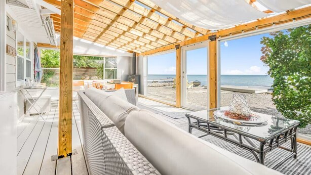A modern beach house with a large wooden deck, a seating area with a sofa and table, and a view of the beach and ocean. - Lake Simcoe Luxury Cottage | Sauna + Hot Tub (Brechin)