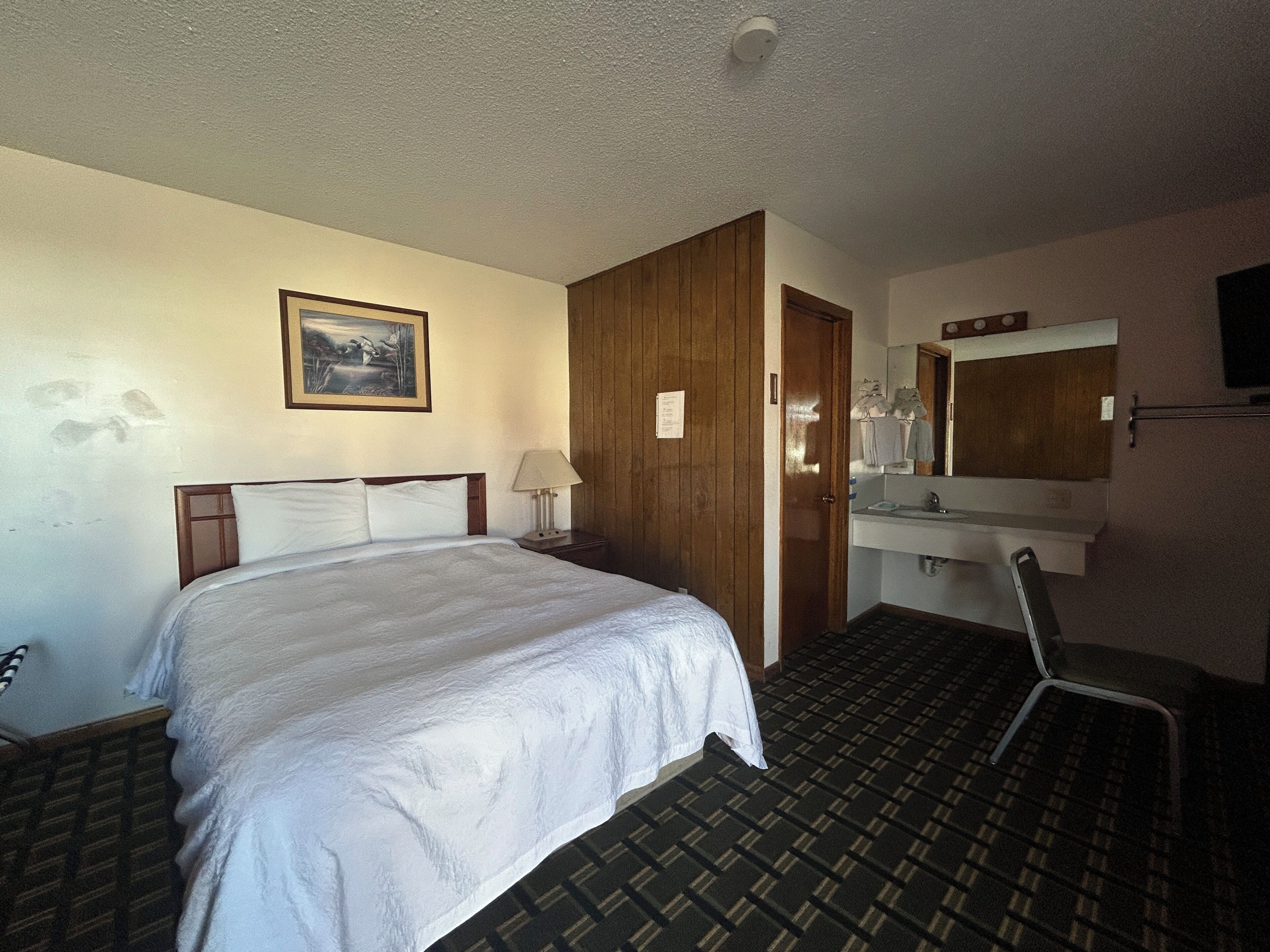 Habitación de hotel con una cama, un escritorio, una silla, un baño con espejo y un cuadro en la pared.