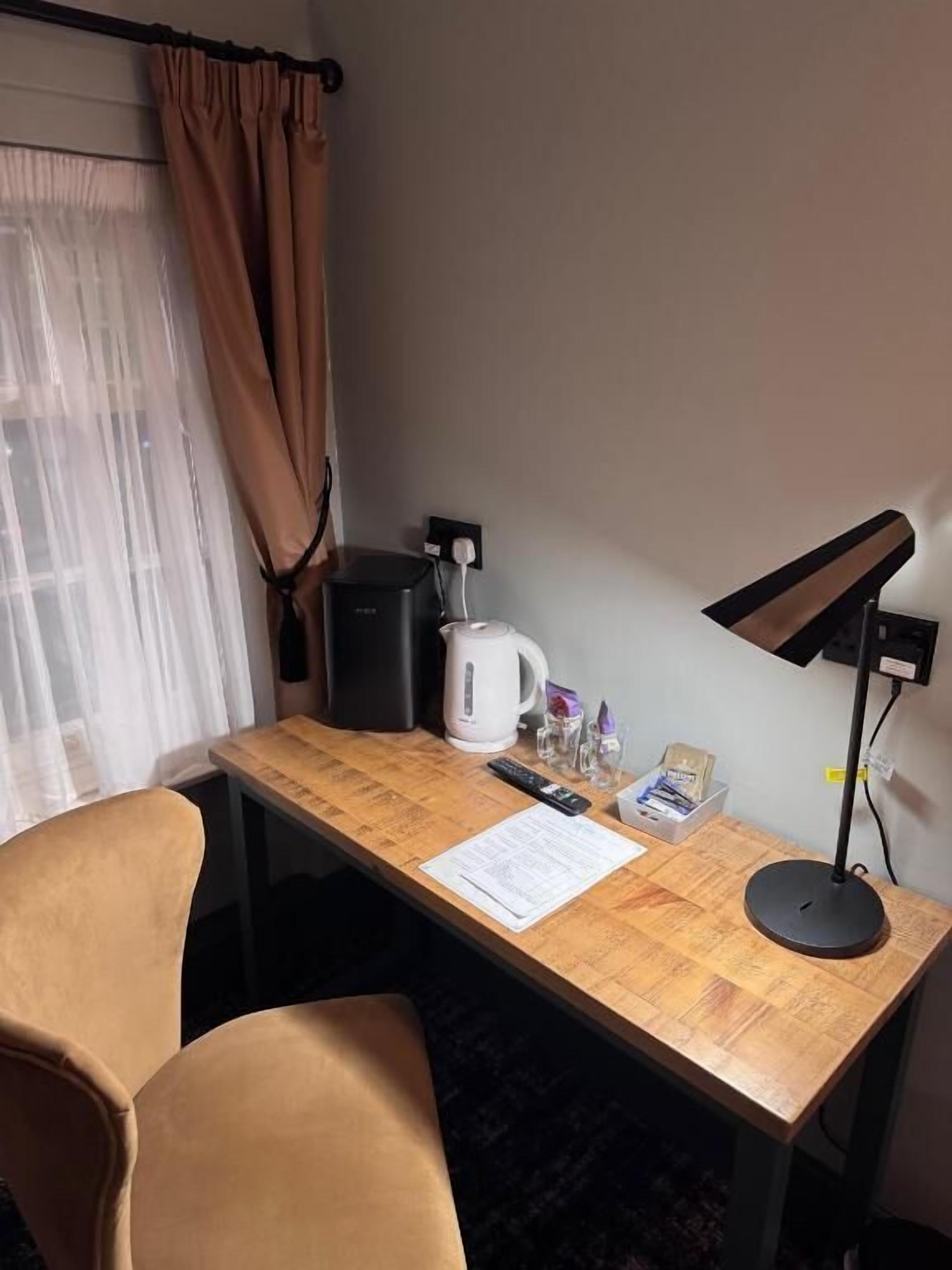 Una camera d'hotel con una scrivania in legno, una lampada da tavolo nera, una macchina del caffè bianca, un telecomando e due sedie.