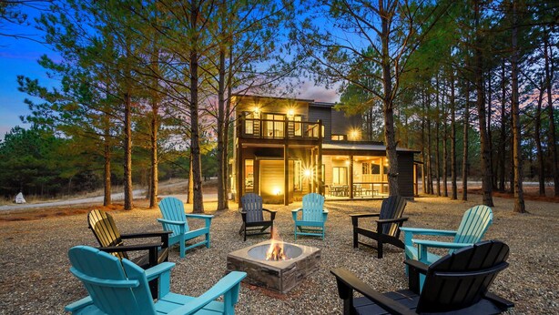 Een modern chalet met twee verdiepingen, met een vuurkuil en zitgedeelte buiten, omringd door bomen.