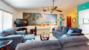 Un salon avec des canapés gris, une table basse en verre, un ventilateur de plafond, un téléviseur et un tableau représentant une scène de plage.