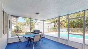 An enclosed patio with a blue carpet, a table with chairs, a grill, and a view of a swimming pool and backyard. - Serenity House — Sunny Private Pool Escape, Dog-Friendly with BBQ Grill & Enclosed Yard 🌞 (Vero Beach)