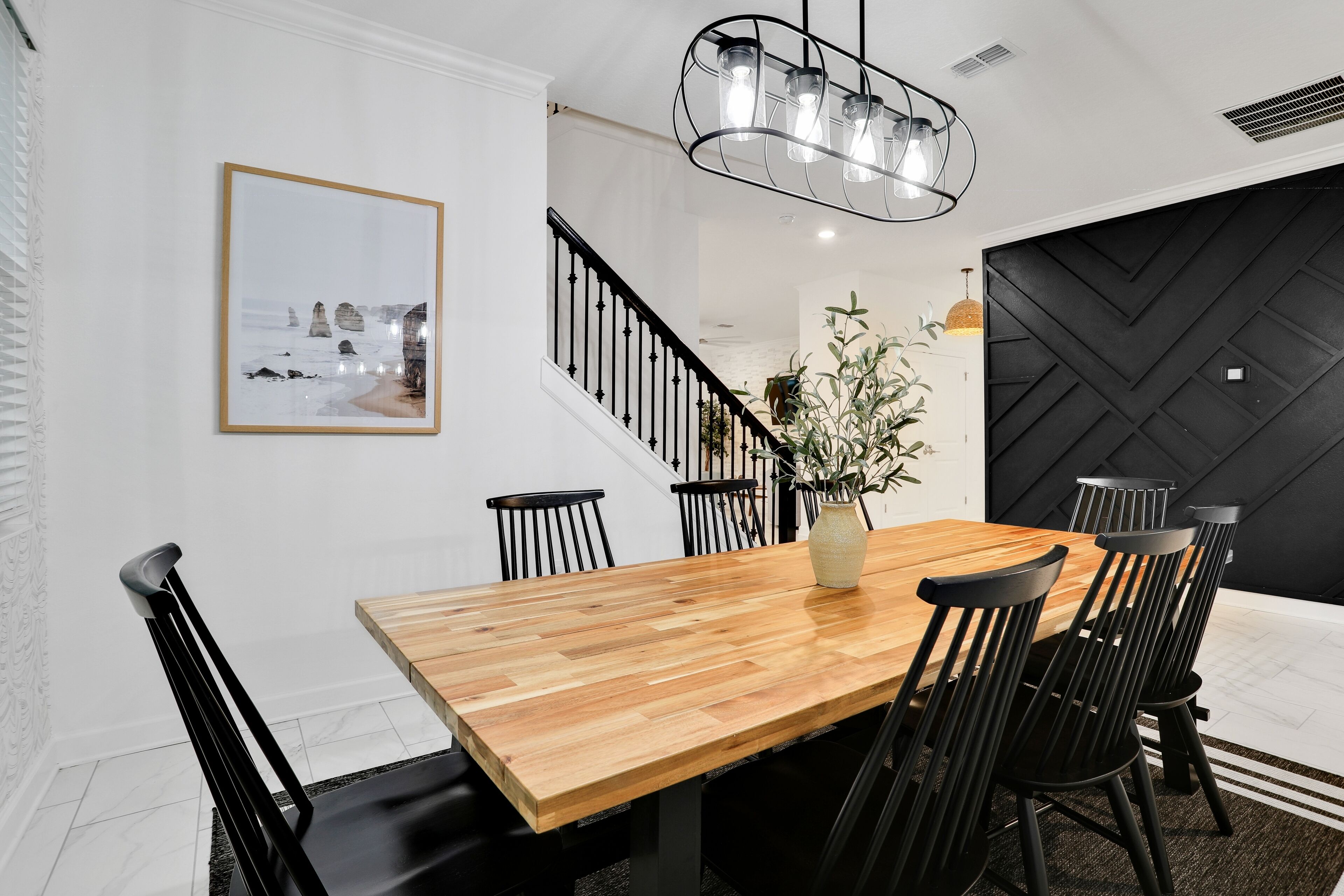 Spiseområde med et træbord, sorte stole, en vase med planter, en trappe og en sort, struktureret væg.