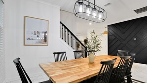 Un espace repas comprenant une table en bois, des chaises noires, un vase avec des plantes, un escalier et un mur noir à texture.