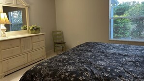 Ein Schlafzimmer mit Bett, Kommode, Stuhl und einem Fenster mit Jalousien.