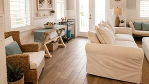 A living room with a wicker sofa, a white armchair, a dining table with a bench, and a view of the outdoors through a glass door. - Gorgeous Home in 30A - Golf Cart + Bikes Included (Santa Rosa Beach)