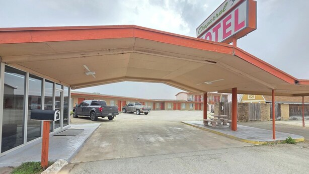 Un motel con área de estacionamiento techada, un letrero y una hilera de habitaciones al fondo.